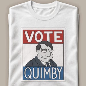 Op de afbeelding: Een wit T-shirt met ronde hals en een rechthoekige grafische print van een stripfiguur in een pak. Het ontwerp bevat de rode tekst VOTE bovenaan en de blauwe tekst QUIMBY onderaan, omlijst door een dunne zwarte rand op de voorkant.