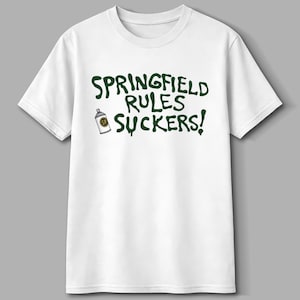 May include: White t-shirt with the text "SPRINGFIELD RULES SUCKERS!" in dark green, accompanied by a small spray paint can graphic. The shirt is a classic crew neck style.