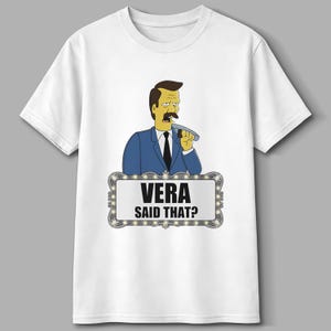 May include: White t-shirt featuring a cartoon illustration of a man in a blue suit holding a plate. The text "VERA SAID THAT?" is displayed on a marquee-style sign below the character. The shirt is a classic crew neck style.