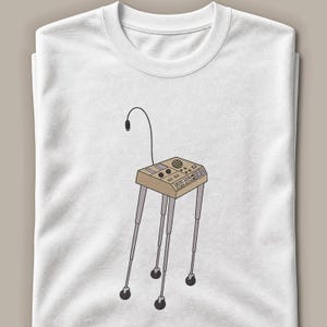 May include: A white t-shirt featuring a graphic illustration of a tan medical monitor or diagnostic device on a tall, four-legged rolling stand. The device includes a small speaker, control knobs, buttons, and a thin black cable extending upwards.