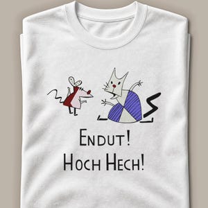 May include: A white crew neck t-shirt featuring a graphic illustration of a small mouse and a larger cat-like creature. Below the characters, the black text reads "ENDUT! HOCH HECH!". The design uses simple black outlines with red and purple accents.