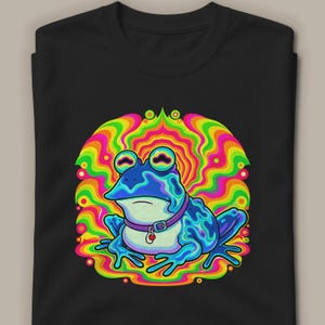 Peut inclure: Un t-shirt noir orné d'une illustration graphique représentant une grenouille bleue portant un collier violet avec une petite étiquette rouge. La grenouille est centrée sur un fond vibrant aux lignes ondulées multicolores, jaunes, orange et vertes.