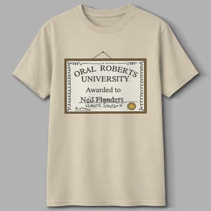 May include: A beige t-shirt featuring a framed certificate design. The certificate reads "Oral Roberts University" and is awarded to "Ned Flanders" with "Homer Simpson" signed below. The design is in black and brown.