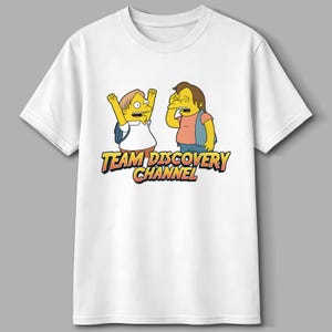 May include: White t-shirt featuring a cartoon graphic of two yellow characters with the text "TEAM DISCOVERY CHANNEL" in orange and yellow. One character is raising their arms, and the other is covering their face.
