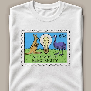 May include: A white t-shirt featuring a graphic of a postage stamp. The stamp shows a tan kangaroo and a purple emu standing on green grass next to a lightbulb. The text reads 60c and 30 YEARS OF ELECTRICITY in black lettering on a blue sky.