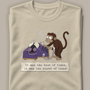 May include: A beige t-shirt featuring a cartoon illustration of a brown monkey with a cigarette, typing on a purple typewriter. A metal chain is attached to the monkey's wrist. Below, a paper graphic reads: It was the best of times, it was the blurst of times!