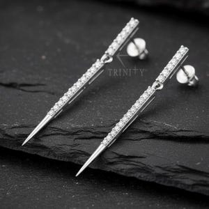 May include: A pair of silver-toned earrings with a linear design. Each earring features a row of small, clear gemstones set along a rectangular bar, tapering to a pointed end. The earrings are displayed on a dark, textured surface.