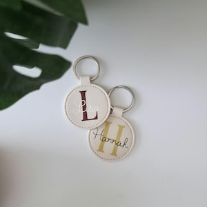 Könnte beinhalten: Zwei cremefarbene runde Schlüsselanhänger mit silbernen Schlüsselringen. Ein Schlüsselanhänger hat den Buchstaben "L" und den Namen "Lisa" in Burgunderrot. Der andere hat den Buchstaben "H" und den Namen "Hannah" in Gold.