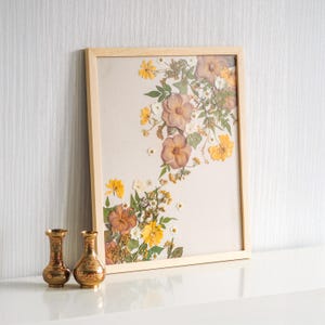 Könnte beinhalten: Ein gerahmtes Kunstwerk mit gepressten Blumen in einem hellen Holzrahmen. Die Anordnung zeigt getrocknete Blumen in Gelb-, Braun- und Weißtönen auf beigem Hintergrund. Zwei kleine, verzierte Goldvasen stehen vor dem gerahmten Kunstwerk.