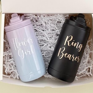 May include: A gift box contains two insulated tumblers. One is a light blue and lavender ombre with "Flower Girl" in white script. The other is black with "Ring Bearer" in gold script. Both tumblers have a matching lid.