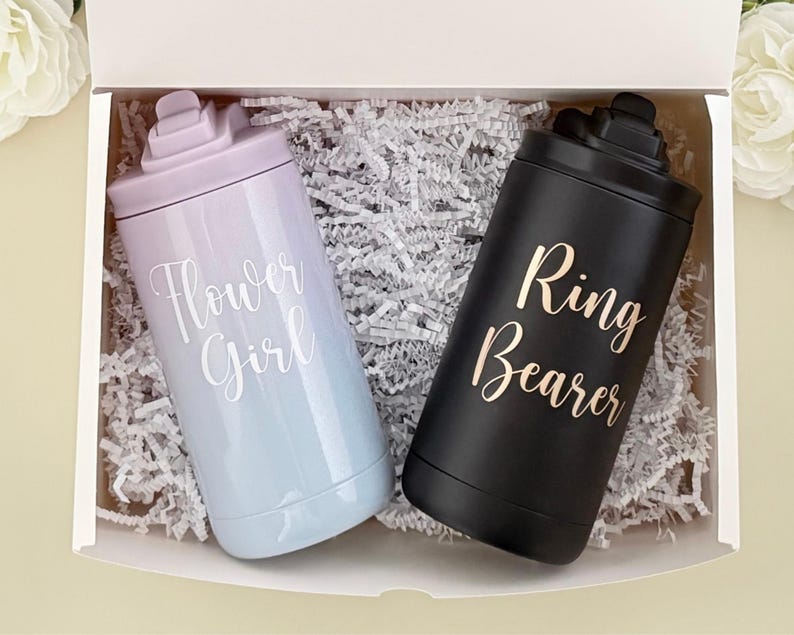 May include: A gift box contains two insulated tumblers. One is a light blue and lavender ombre with "Flower Girl" in white script. The other is black with "Ring Bearer" in gold script. Both tumblers have a matching lid.