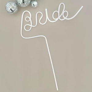May include: A white, cursive "Bride" cake topper with a long stem, perfect for a wedding cake. The topper is accompanied by three silver disco ball ornaments. The design is simple and elegant, suitable for a bridal shower or wedding.