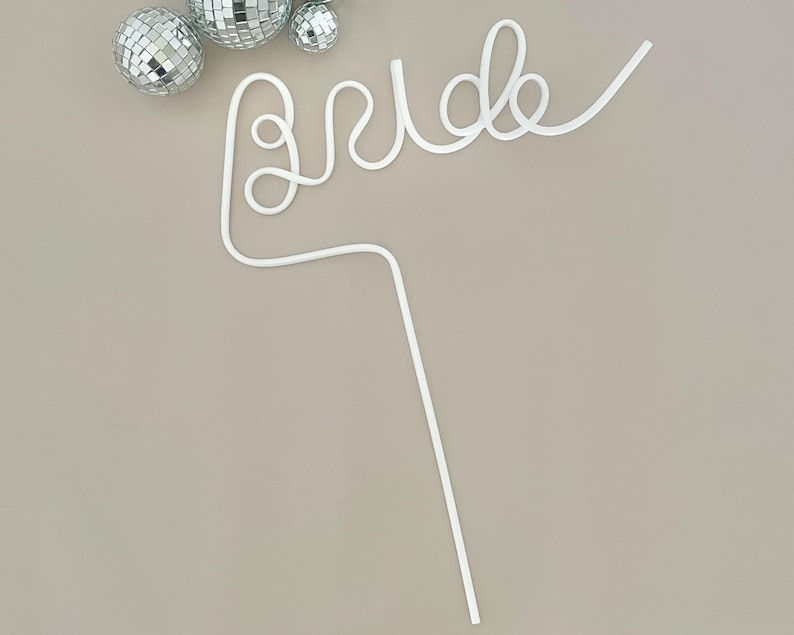 May include: A white, cursive "Bride" cake topper with a long stem, perfect for a wedding cake. The topper is accompanied by three silver disco ball ornaments. The design is simple and elegant, suitable for a bridal shower or wedding.