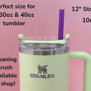 May include: A green 30oz or 40oz Stanley tumbler with a lid and a handle. The tumbler has a silver band around the middle and the Stanley logo is visible. A purple 12-inch straw with glitter is inserted into the tumbler.