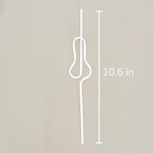 May include: A white, decorative metal hair pin, measuring 10.6 inches in length. The pin has a unique, abstract design with a looped central section. The background is a neutral beige color.