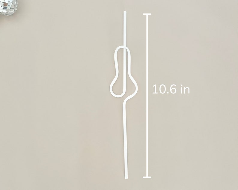 May include: A white, decorative metal hair pin, measuring 10.6 inches in length. The pin has a unique, abstract design with a looped central section. The background is a neutral beige color.