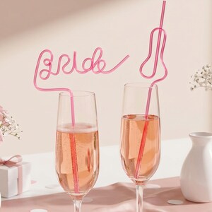 May include: Two champagne flutes filled with a pink beverage, each with a pink straw. One straw spells "Bride". The glasses are on a pink surface, with pink roses and a gift box in the background. A celebratory theme.