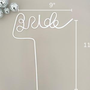May include: White wire cake topper that spells "Bride", measuring 9 inches wide and 11 inches tall. The topper is accompanied by several silver disco balls. Perfect for a bridal shower or wedding.