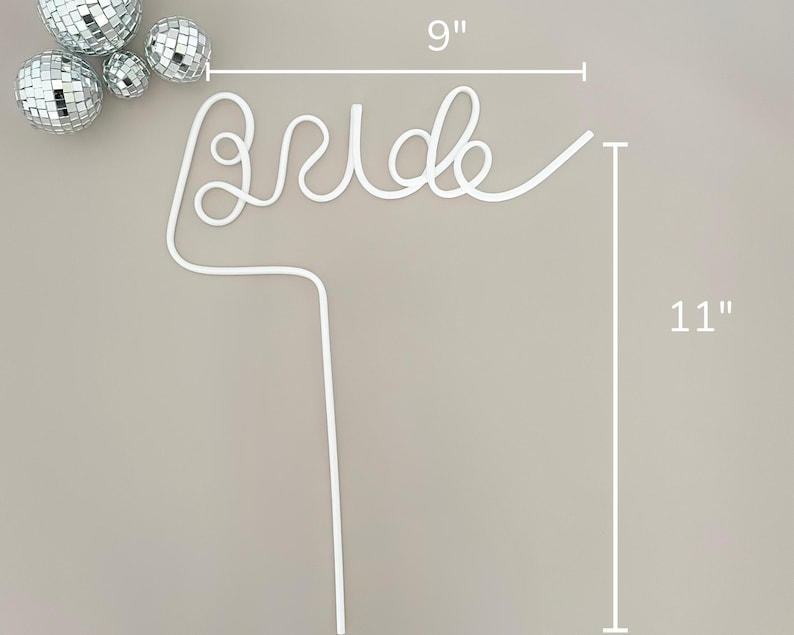May include: White wire cake topper that spells "Bride", measuring 9 inches wide and 11 inches tall. The topper is accompanied by several silver disco balls. Perfect for a bridal shower or wedding.