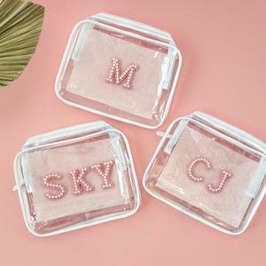 May include: Three clear plastic zippered pouches with white trim. Each pouch has a different letter or name spelled out in pink pearl beads. The pouches are on a pink background.