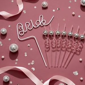 May include: A white wire cake topper featuring the word bride in cursive script, accompanied by eight clear plastic spiral straws, each topped with a small silver disco ball, arranged on a dark pink surface with scattered silver confetti.