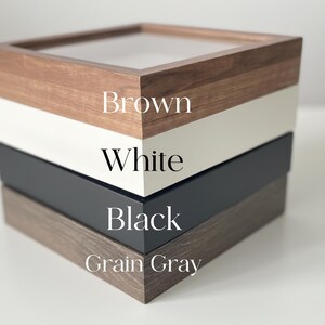 May include: A square wooden box with a clear top, featuring layers of brown, white, black, and grain gray. The text on the side of the box indicates the color of each layer.