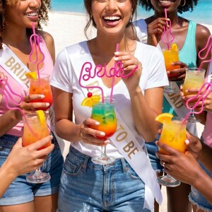 May include: A group of people holding colorful cocktails with pink decorative straws. The cocktails have orange and yellow garnishes. Some people are wearing white sashes that say "Bride to Be".