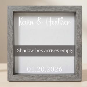 May include: A gray wooden shadow box frame with white text that reads "Kevin & Heather", "Shadow box arrives empty", and the date "01.20.2026". The frame has a light gray background.