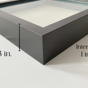 May include: A black shadow box frame with a glass front. The frame is 1.8 inches deep, with an interior depth of 1 inch. The text "Interior 1 in." is visible on the right side of the frame.