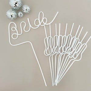 May include: White plastic straws shaped to spell the word "bride" with additional straws in a bundle below. The straws are on a light brown surface with silver disco balls scattered around.