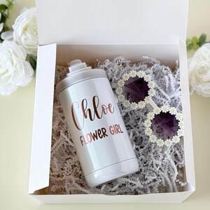 May include: A white gift box contains a white tumbler with "Chloe FLOWER GIRL" in rose gold script, and sunglasses with daisy-shaped frames. The box is filled with white shredded paper.
