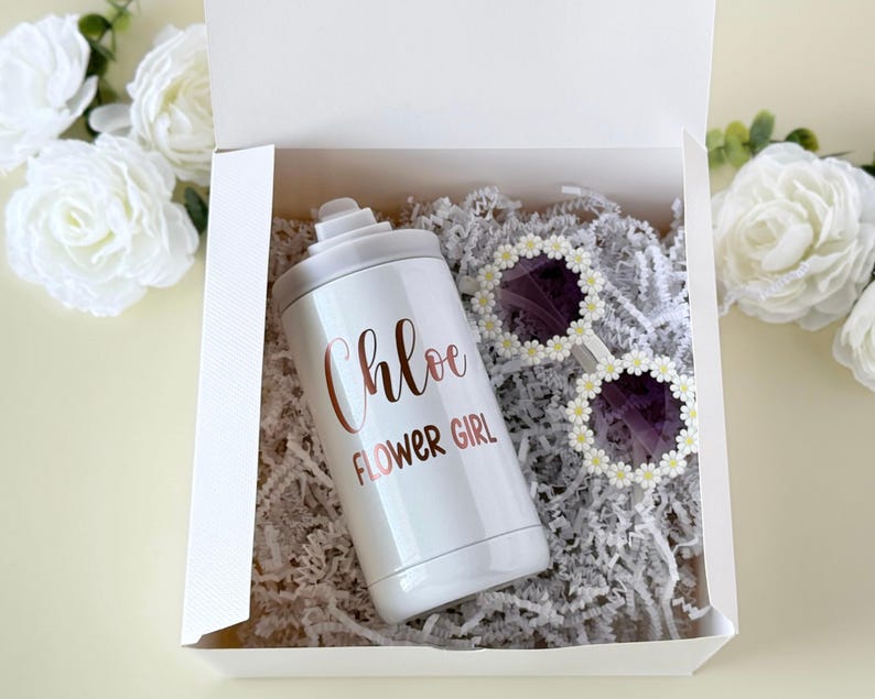 May include: A white gift box contains a white tumbler with "Chloe FLOWER GIRL" in rose gold script, and sunglasses with daisy-shaped frames. The box is filled with white shredded paper.