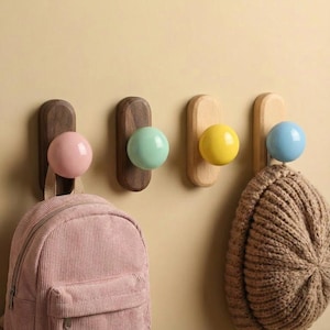 Op de afbeelding: Vier houten wandhaken met gekleurde bolvormige knoppen. De haken zijn aan een lichtgekleurde muur bevestigd. Een roze corduroy rugzak en een bruine gebreide muts hangen aan twee van de haken. De knoppen zijn roze, groen, geel en blauw.