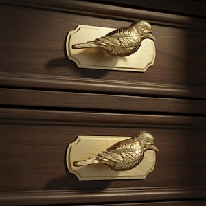 May include: Two brass bird-shaped drawer pulls mounted on rectangular backplates. The drawer pulls are gold-colored and detailed, with the birds facing right. The background is a dark brown wooden surface.