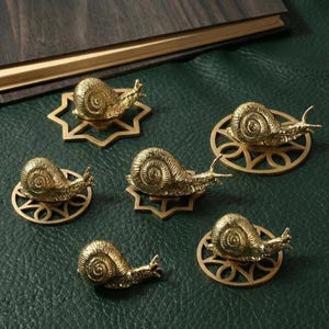 Op de afbeelding: Zes messing slakkenbeeldjes, elk met gedetailleerde schelp- en lichaamskenmerken. De slakken rusten op decoratieve messing bases, sommige met stervormige ontwerpen, andere met cirkelvormige patronen. Gerangschikt op een donkergroen oppervlak.