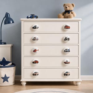 May include: A white five-drawer dresser featuring round knobs decorated with colorful illustrations of emergency vehicles, including a police car, fire truck, ambulance, motorcycle, helicopter, tow truck, SUV, boat, and rescue van.