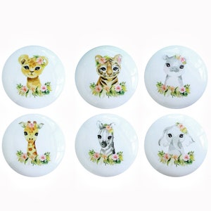 Może przedstawiać: Sześć okrągłych ceramicznych gałek z różnymi twarzami zwierząt. Każda gałka przedstawia inne zwierzę, w tym lwa, tygrysa, zebrę, żyrafę, słonia i hipopotama. Każde zwierzę nosi wianek z kwiatów i jest otoczone zielonymi liśćmi i różowymi kwiatami.