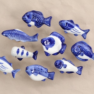 May include: Nine ceramic fish ornaments in blue and white. The fish vary in design, with detailed scales and fins. They are arranged on a neutral-coloured surface, offering a decorative nautical theme.