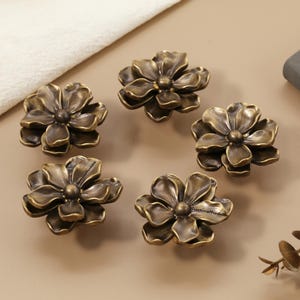 May include: Five antique brass flower-shaped cabinet knobs. Each knob has detailed petal designs and a central, rounded core. The knobs are arranged on a neutral surface, suggesting a decorative hardware theme.