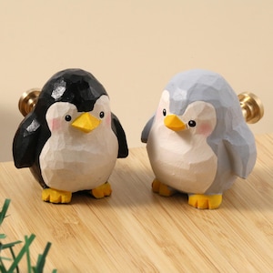 May include: Two wooden penguin cabinet knobs. One is black and white, the other is gray and white. Both have yellow beaks and feet, and pink cheeks. The knobs have a gold-colored metal screw.