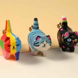 May include: Three hand-painted wooden cat figurines. One is rainbow-colored, another is blue with stripes, and the third is black with pink polka dots. Each cat has a gold-colored ring attached to its back.