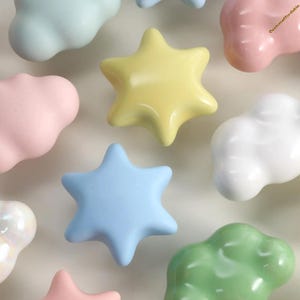 May include: A collection of pastel-coloured ceramic decorative knobs in cloud and star shapes. Colours include light blue, pink, yellow, green, and white. These knobs are likely for furniture or decorative projects.