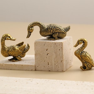 Pode incluir: Três estatuetas de pato de latão antigo. Um pato está sobre um pequeno bloco retangular, outro sobre um bloco quadrado e o terceiro sobre uma superfície plana. Os patos têm texturas de penas detalhadas e estão em várias poses.