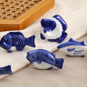 May include: A selection of ceramic fish ornaments in blue and white. The fish have different designs, some with scales and others with smooth surfaces. These decorative items are displayed on a white cloth.