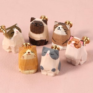 Puede incluir: Seis figuras de gato de madera talladas a mano con detalles de latón. Cada gato está pintado con diferentes colores y patrones, incluyendo marrón, blanco, naranja y gris. Los gatos están colocados sobre una superficie rosa.