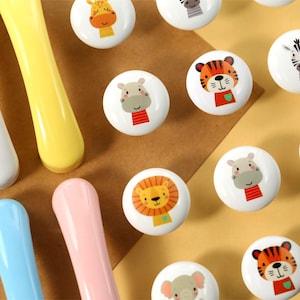 Könnte beinhalten: Sortiment an weißen Keramik-Schubladengriffen mit bunten Tierillustrationen. Die Tiere umfassen eine Giraffe, ein Zebra, einen Tiger, einen Löwen, ein Flusspferd und einen Elefanten. Die Griffe werden auf einer braunen und gelben Oberfläche präsentiert.