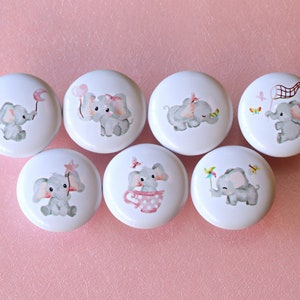Puede incluir: Conjunto de seis pomos de cajón de cerámica blanca con lindas ilustraciones de elefantes grises. Cada pomo presenta un diseño de elefante diferente con acentos rosados ​​y detalles como un globo, una mariposa, una taza, una estrella, una red y un molinete.