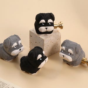 May include: Four wooden dog figurines, two in black and two in grey, with brass-coloured hardware. One black dog sits atop a small, square stone block. The dogs have white accents on their faces and chests.