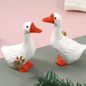 Könnte beinhalten: Zwei weiße, gänseförmige Schrankknöpfe mit orangefarbenen Schnäbeln und Füßen. Die Knöpfe haben eine goldfarbene Basis. Die Gänse befinden sich auf einer grauen Oberfläche mit rosa Hintergrund.