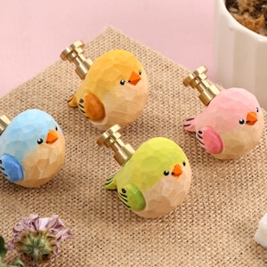 May include: Four colourful, carved wooden bird figurines with brass-coloured stems. The birds are in shades of blue, yellow, green, and pink, each with an orange beak and black eyes. They rest on a textured, tan surface.